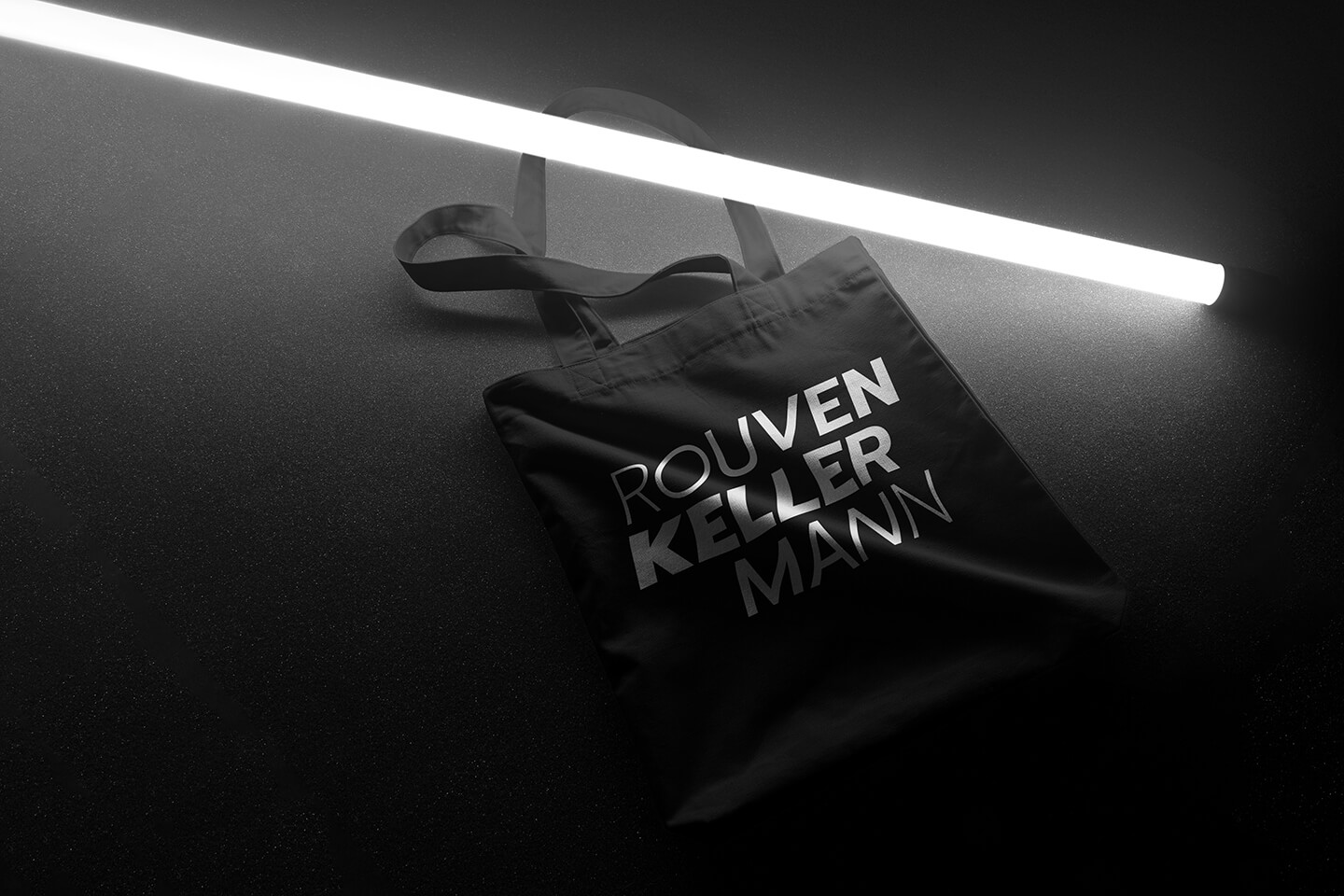Schwarze Rouven Kellermann Tragetasche mit weißem Logo unter einer hellen Leuchtstoffröhre.